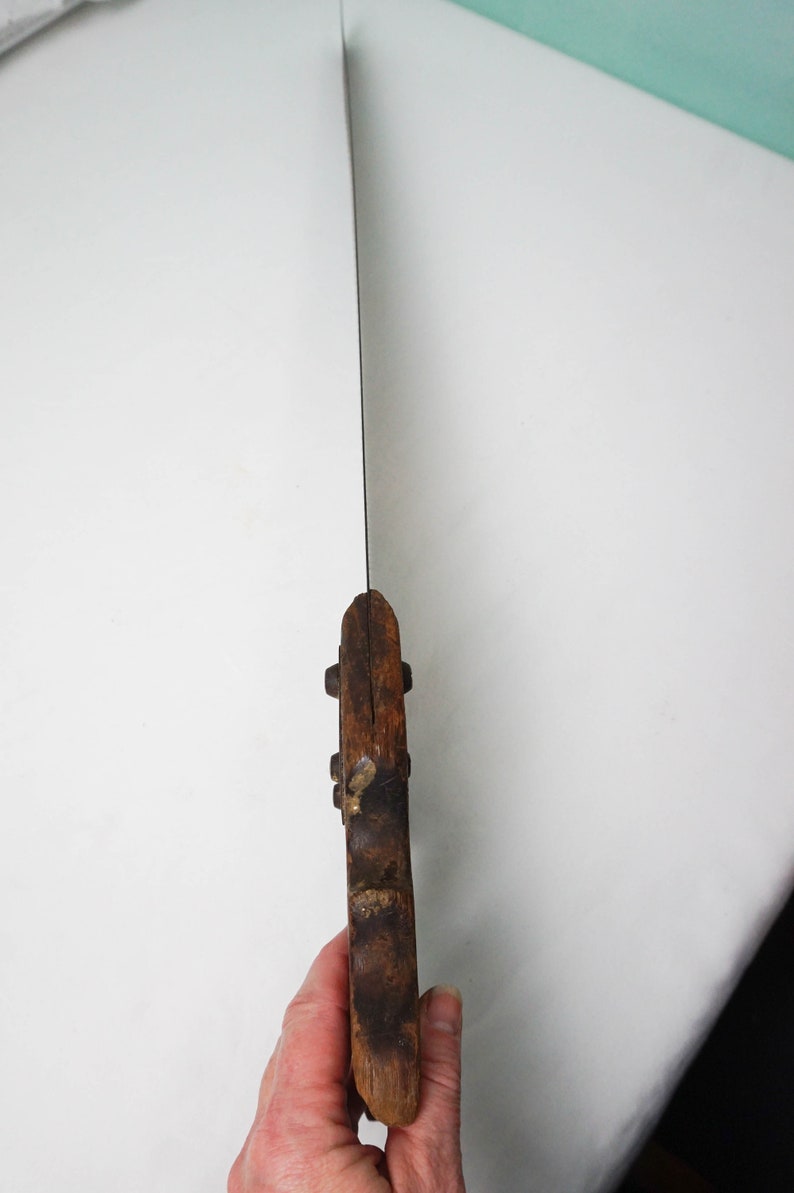 May include: A vintage hand saw with a wooden handle. The saw blade is made of metal and has a slight curve. The handle is brown and has a few small metal rivets.