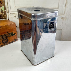 May include: A vintage chrome flour canister with a black handle on top. The rectangular container has a small label that reads "Flour" with a graphic of a stack of flour sacks. The canister is reflective and sits on a white surface.