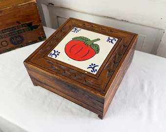 May include: A square wooden box with a decorative tile top featuring a red tomato design with green leaves. The box is made of dark brown wood with carved details around the edges and is elevated on small feet.