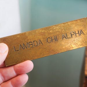 Pode incluir: Uma placa de metal retangular, cor de lat&atilde;o, com o texto gravado "LAMBDA CHI ALPHA". A placa tem um pequeno orif&iacute;cio numa extremidade. A placa &eacute; segurada em uma m&atilde;o.