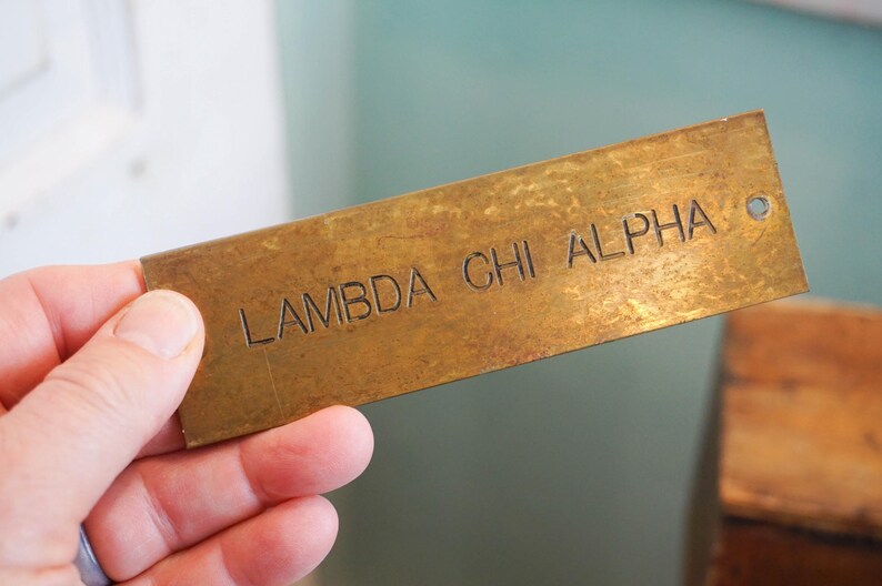 Pode incluir: Uma placa de metal retangular, cor de lat&atilde;o, com o texto gravado "LAMBDA CHI ALPHA". A placa tem um pequeno orif&iacute;cio numa extremidade. A placa &eacute; segurada em uma m&atilde;o.
