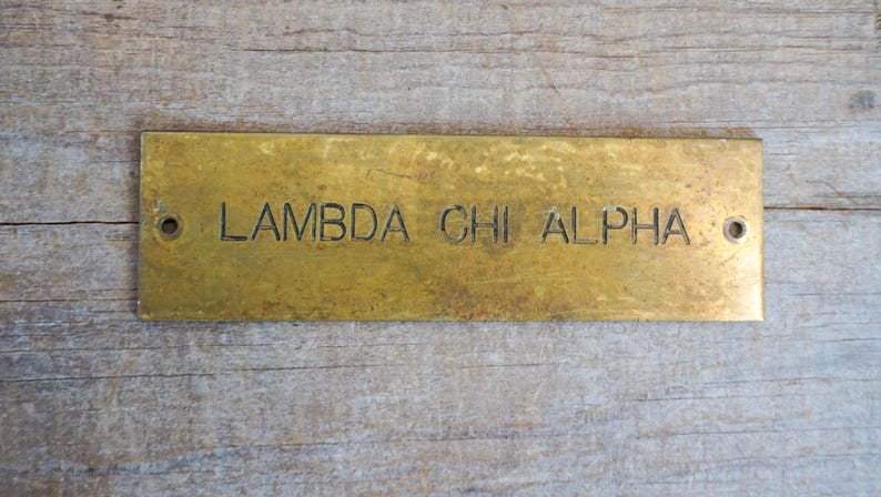 Pode incluir: Placa retangular de lat&atilde;o com o texto gravado "LAMBDA CHI ALPHA". A placa tem dois orif&iacute;cios, um em cada extremidade. O lat&atilde;o tem uma apar&ecirc;ncia desgastada e envelhecida. A placa est&aacute; sobre um fundo de madeira.