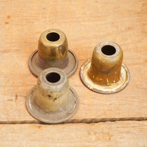May include: Three vintage brass lamp parts with a circular base and a cylindrical top. The metal has a tarnished, aged appearance, with variations in color from gold to bronze. The objects are arranged on a wooden surface.