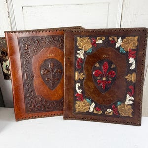 May include: Two brown leather journals with embossed designs. One features a fleur-de-lis and the initials "G.C.R.", the other has a fleur-de-lis with red, white, and gold accents and the initials "P.L.L.".