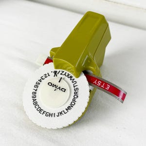 May include: A vintage olive green Dymo embossing label maker with a white circular dial featuring black letters and numbers. A red adhesive strip is protruding from the side of the device, clearly displaying the embossed white text reading ETSY.