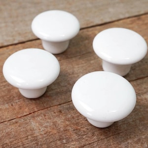 May include: Four white ceramic cabinet knobs with a rounded top and a short stem. The knobs are arranged in a semi-circle on a wooden surface.