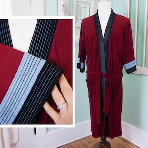 May include: A burgundy robe with black and light blue striped trim on the lapels, cuffs, and belt. The robe has a shawl collar, pockets, and a matching belt. The robe is displayed on a mannequin.