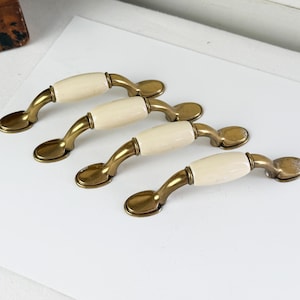 May include: Four cabinet pulls with a brass finish and cream-colored ceramic center. The pulls are curved with a classic design, suitable for drawers or cabinet doors. The hardware is arranged on a white surface.