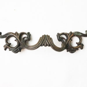 May include: A vintage ornate metal drawer pull with a decorative, scrolled design. The pull is finished in a dark bronze color.