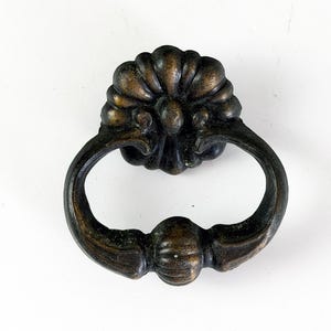 May include: Antique bronze-toned drawer pull with a decorative floral design and a ring handle. The hardware is likely made of metal and is designed for furniture. The pull is set against a white background.
