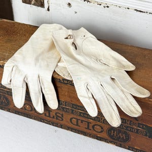 May include: A pair of off-white leather gloves featuring a smooth texture and a small, circular metallic button closure at the wrist. The gloves are designed with stitched finger seams and a classic cut, showing signs of wear on the surface.
