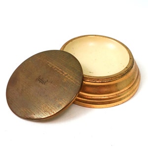 May include: A small, round, gold-colored metal trinket box with a lid. The lid has a textured surface and the letter "F" engraved on it. The box has a white interior.
