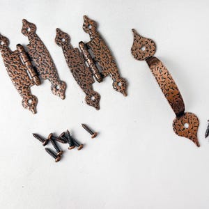 May include: Two copper-colored metal hinges with a textured finish and decorative pointed ends, accompanied by a matching arched cabinet handle and a collection of small black metal screws for installation on furniture or cabinetry hardware.