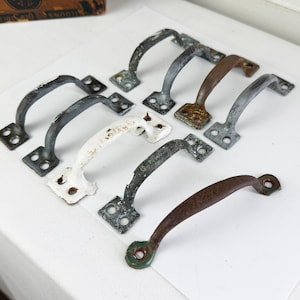 May include: A collection of nine antique metal drawer pulls in various colors, including black, white, and brown. Each pull has a curved handle and a rectangular base with screw holes. The pulls are arranged on a white surface.