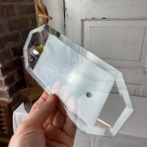 May include: A clear, geometric-shaped glass object with a beveled edge is held in a hand. The object has a rectangular shape with angled sides and a small hole. The background includes a white door and a brick wall.