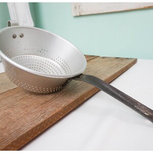 May include: A silver aluminum colander with a black metal handle. The colander has a round bowl shape and a perforated bottom. It is sitting on a wooden surface.