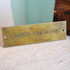 Pode incluir: Placa retangular de lat&atilde;o com o texto gravado "LAMBDA CHI ALPHA". A placa tem uma apar&ecirc;ncia desgastada e envelhecida com dois pequenos orif&iacute;cios. Uma caixa de madeira &eacute; vis&iacute;vel ao fundo.