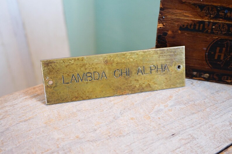 Pode incluir: Placa retangular de lat&atilde;o com o texto gravado "LAMBDA CHI ALPHA". A placa tem uma apar&ecirc;ncia desgastada e envelhecida com dois pequenos orif&iacute;cios. Uma caixa de madeira &eacute; vis&iacute;vel ao fundo.