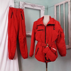 May include: A red ski suit with a black zipper and belt. The jacket has a hood and a pocket on the left sleeve. The pants have a black zipper on the side.