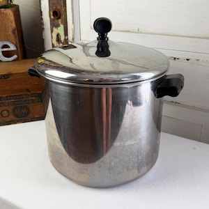 May include: A stainless steel stockpot with a polished finish, featuring two black side handles and a matching lid with a black knob. The cylindrical pot has a reflective surface and a flat base, designed for cooking large quantities of food.