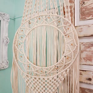 May include: A large, cream-colored macrame wall hanging with a circular design and wooden dowel at the top. The macrame has a fringe of tassels and small, brown beads.