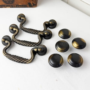 May include: Three antique brass-finished metal drawer pulls with a twisted rope design and four matching round cabinet knobs. Each piece features a dark, weathered patina with highlighted gold-toned edges, providing a textured appearance.