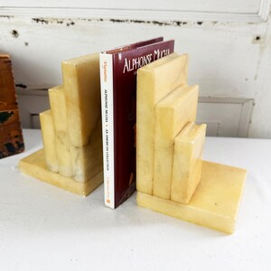 May include: A pair of cream-colored, polished stone bookends with a stepped, tiered design, each featuring a smooth, marbled texture. The bookends are positioned on either side of a single book titled Alphonse Mucha: An American Collection.