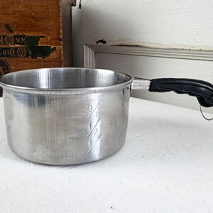 May include: A small, brushed aluminum saucepan with a black handle and a metal loop. The interior of the pan has measurement markings for 1/2, 3/4, and 1 cup. The pan is designed for cooking and food preparation.