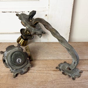 May include: Antique door hardware set, including a decorative handle with a twisted design, a doorknob, and a lockset. The metal pieces have a weathered, aged appearance, with a mix of silver and brass tones. The set is displayed on a wooden surface.