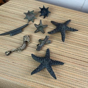 May include: An assortment of decorative metal objects, including starfish, seahorse, and coral shapes, in shades of black and bronze. The objects are arranged on a light brown woven surface.
