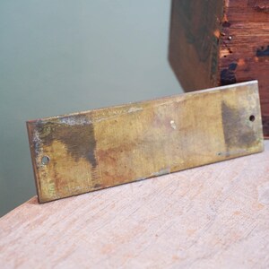 Pode incluir: Uma placa retangular de lat&atilde;o com dois furos, mostrando sinais de idade e desgaste. A placa tem um tom dourado manchado com manchas mais escuras. Uma caixa de madeira &eacute; parcialmente vis&iacute;vel no fundo.