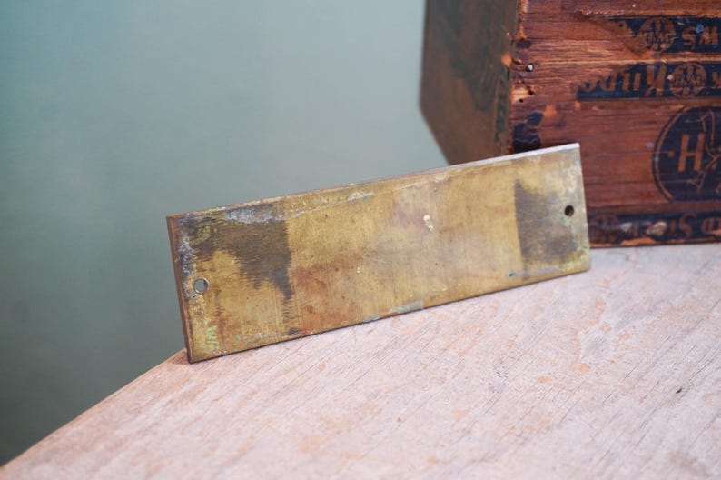 Pode incluir: Uma placa retangular de lat&atilde;o com dois furos, mostrando sinais de idade e desgaste. A placa tem um tom dourado manchado com manchas mais escuras. Uma caixa de madeira &eacute; parcialmente vis&iacute;vel no fundo.