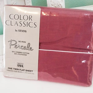 May include: A pink twin flat sheet made of 50% combed cotton and 50% polyester. The sheet is packaged in plastic and has a white label with the text "Color Classics by Stevens, No Iron Percale, Made in the USA, One Twin Flat Sheet, Finished Size 39" x 75" (99 x 191 cm)".