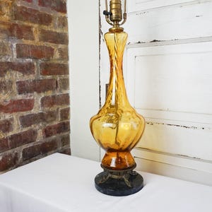May include: A vintage amber glass table lamp with a unique, elongated shape. The lamp features a brass neck and a dark, decorative base. The lamp is placed on a white surface, with a brick wall and white door in the background.