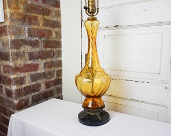 May include: A vintage amber glass table lamp with a unique, elongated shape. The lamp features a brass neck and a dark, decorative base. The lamp is placed on a white surface, with a brick wall and white door in the background.