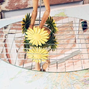 May include: A decorative mirror with a grid pattern, reflecting a yellow flower arrangement and a wooden mannequin. The mirror is oval-shaped and sits on a map, creating a unique display.