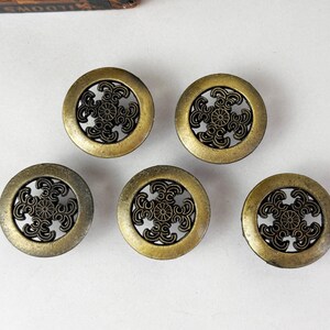 May include: Five circular brass-toned metal buttons featuring an openwork design. Each button has a solid outer ring and a central motif composed of four symmetrical, curved scroll-like elements radiating from a small, detailed circular center.