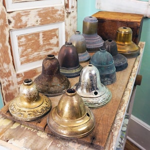 May include: A collection of vintage metal lamp shades in various shapes and finishes, including brass, copper, and silver. The shades are displayed on a weathered wooden surface, showcasing their aged patina and unique designs. The collection includes shades with decorative details.