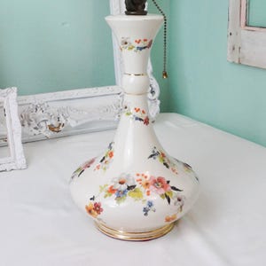 May include: A white ceramic table lamp with a floral pattern in pink, orange, blue, and yellow. The lamp has a bulbous base and a slender neck, with gold trim at the base. A pull chain hangs from the top.