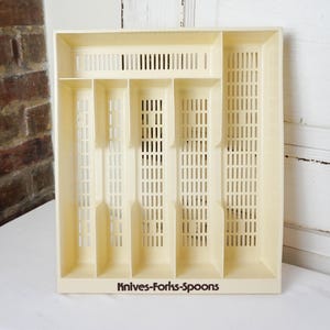 May include: A cream-colored plastic cutlery organizer with multiple compartments. The organizer has a rectangular shape with slotted ventilation and the text "Knives-Forks-Spoons" printed on the front.