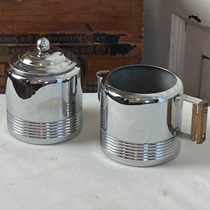 May include: A chrome-plated sugar bowl with a lid and a creamer with a wooden handle. Both pieces have horizontal ribbed detailing around the base. The set is displayed on a white surface.