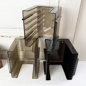 May include: Three translucent, dark-tinted plastic storage racks for media or documents. Each unit features a U-shaped frame with multiple horizontal slots along the interior sides for organizing items in a vertical, stacked arrangement.