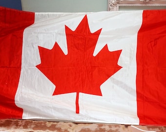 Pode incluir: Bandeira canadense retangular com uma folha de bordo vermelha no centro. A bandeira tem faixas vermelhas verticais nas laterais e uma faixa branca no meio. O tecido é leve.