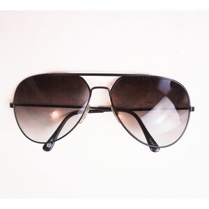 May include: Black aviator sunglasses with gradient lenses. The sunglasses have a thin black metal frame and a double bridge. The lenses transition from dark brown at the top to a lighter shade at the bottom. The sunglasses are set against a white background.