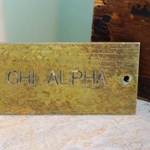 Pode incluir: Uma placa de lat&atilde;o desgastada com o texto gravado "BDA CHI ALPHA". A placa tem um &uacute;nico furo e est&aacute; colocada sobre um fundo desfocado. Um objeto de madeira &eacute; parcialmente vis&iacute;vel.