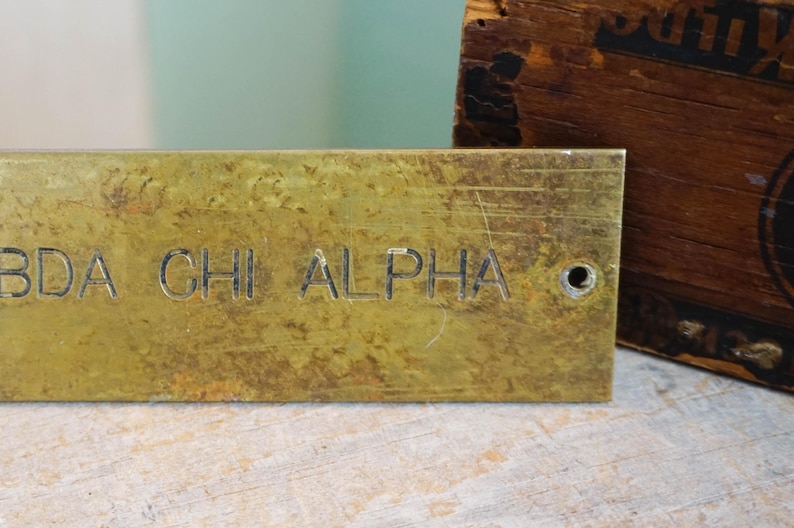 Pode incluir: Uma placa de lat&atilde;o desgastada com o texto gravado "BDA CHI ALPHA". A placa tem um &uacute;nico furo e est&aacute; colocada sobre um fundo desfocado. Um objeto de madeira &eacute; parcialmente vis&iacute;vel.