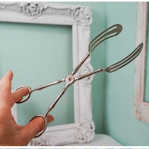 May include: Silver-toned serving tongs with a fork-like design on one end and a ring on the other. The tongs are held in a hand against a light blue background.