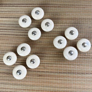 May include: A collection of eleven off-white ceramic cabinet knobs with speckled detailing. Each knob features a silver-colored metal center. The knobs are arranged on a light brown woven surface.