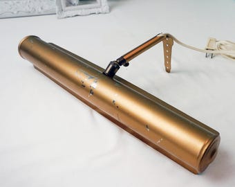 May include: A vintage brass-toned picture light with an adjustable arm and a plug-in cord. The light fixture has a cylindrical body with some paint wear, suggesting age. The adjustable arm is connected to a mounting bracket.