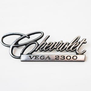 May include: A vintage Chevrolet Vega 2300 emblem. The emblem features the Chevrolet script in a cursive font above a rectangular bar with the text "VEGA 2300". The emblem is silver and black.